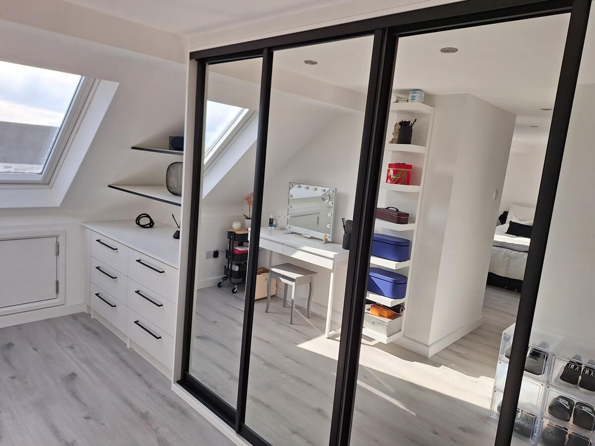 Full-mirror sliding wardrobes, open shelving and a dressing table, fitted into a loft conversion dressing area with a skylight
