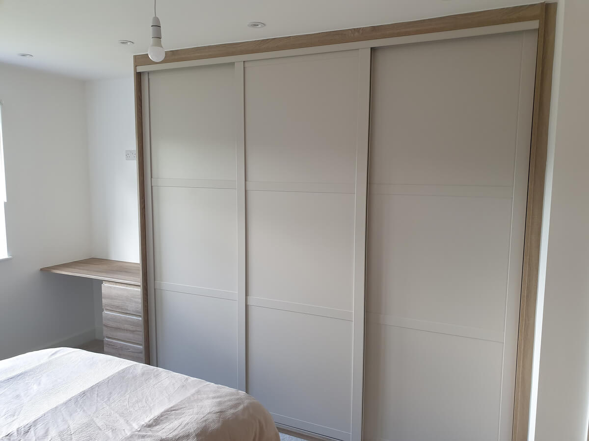 Three Cashmere SHAKER sliding wardrobe doors in an oak-effect surround with a built-in desk and drawers, fitted in a bedroom in Stafford
