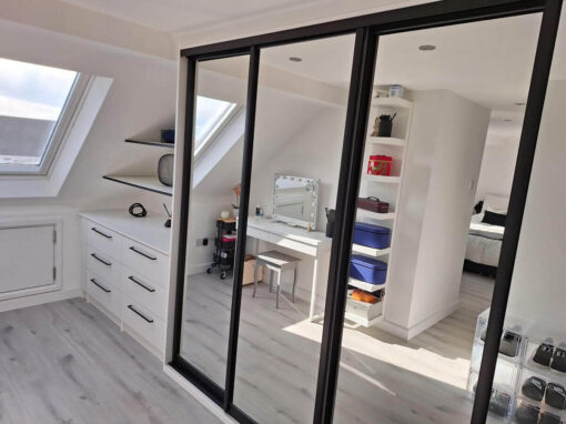 Black-framed mirror sliding wardrobe doors in a loft dressing area with a dressing table and drawers under the skylight